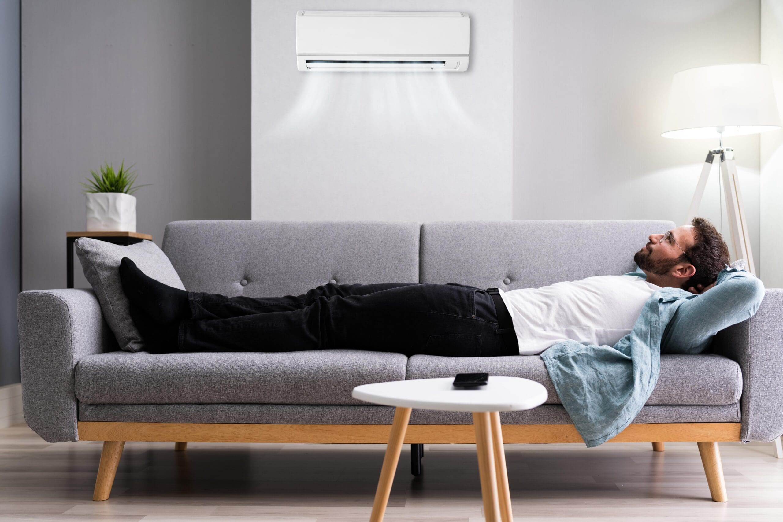 Man relaxing on a gray sofa in a well-lit living room with an air conditioning unit above, illustrating comfort during summer heat while discussing HVAC issues.