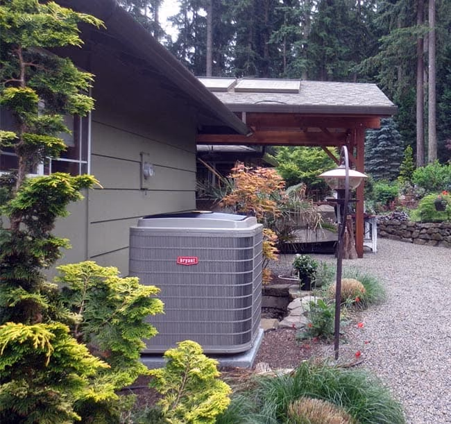 Air conditioner condenser beside a home with evergreens and shrubs