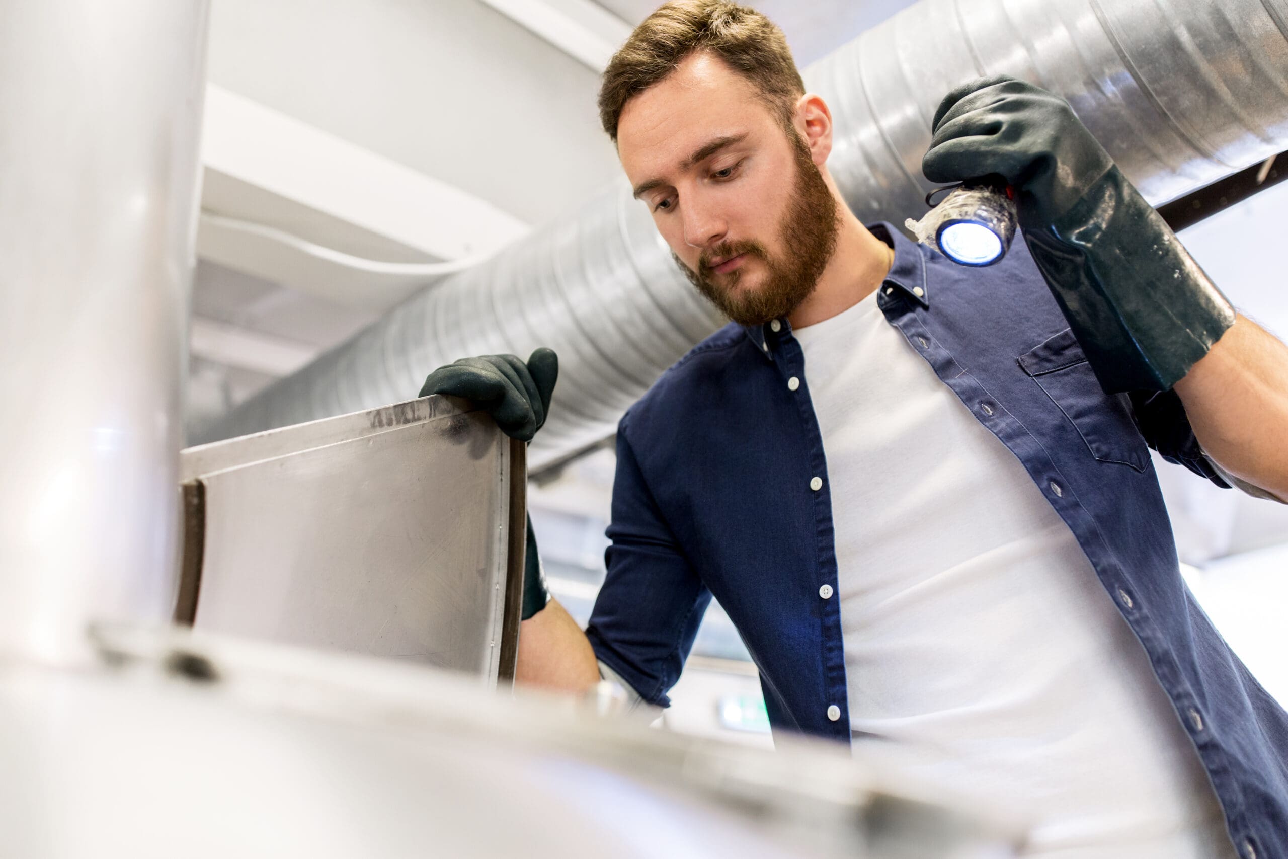 What is undersized return air duct in Portland? Man inspecting equipment in a brewery, wearing gloves and holding a flashlight, with ductwork visible in the background, emphasizing HVAC system maintenance.